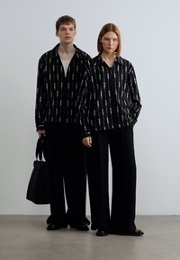 Δύο μοντέλα φορούν ταιριαστές μαύρες και λευκές ριγέ μπλούζες με μακριά μανίκια, συνδυασμένες με φαρδιά μαύρα παντελόνια. Το ένα κρατάει μια μαύρη τσάντα.