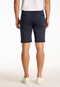 Navy blue shorts with a smooth texture, two back pockets with buttons, hemmed edges, and a fitted design, paired with white sneakers.