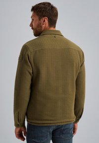 Mężczyzna odwrócony tyłem, ubrany w fakturowaną oliwkowozieloną kurtkę z zapiętymi mankietami i naszywką na lewym rękawie, w zestawieniu z niebieskimi dżinsami.