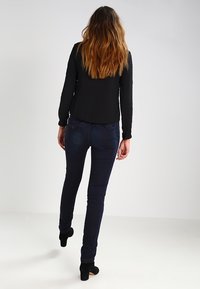 Haut noir à manches longues associé à un jean skinny bleu foncé et des bottines noires. Tissu lisse et design ajusté. Cheveux tombant le long du dos.
