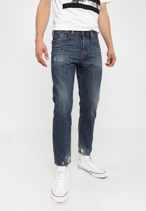 Mann trägt ausgebleichte blaue Jeans mit ausgefransten Saumkanten und weiße High-Top-Sneaker, steht vor einem einfarbigen Hintergrund.