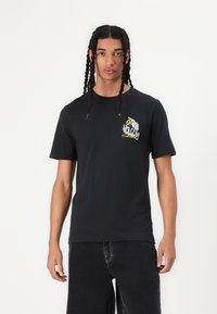 Black cotton t-shirt featuring a colorful graphic design on the left chest. Short sleeves and crew neck. Model wears casual dark denim pants.