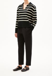 Personne portant un pantalon noir court, un haut à rayures noir et blanc à manches longues en V avec col, et des chaussures plates noires avec des brides de cheville.