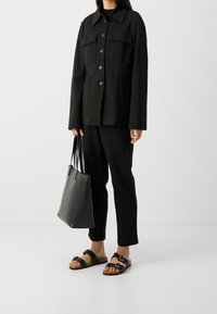 Czarna kurtka użytkowa z dużymi kieszeniami na piersi, zestawiona z czarnymi spodniami i skórzanymi sandałami. Całość uzupełnia usztywniana czarna torba typu tote.