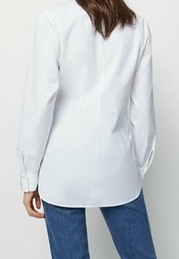 Blouse blanche à manches longues avec une texture lisse, dotée d'un ourlet incurvé et de poignets boutonnés. Détail de couture centrale visible le long de la colonne vertébrale.