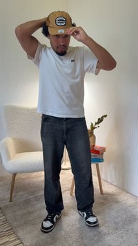 Brūna cepure ar sietveida aizmuguri un logotipu, balta T-krekls, tumšas džinsa bikses, melni-baltas sporta kurpes. Iekštelpu vide ar maigām tekstūrām un siltu apgaismojumu.