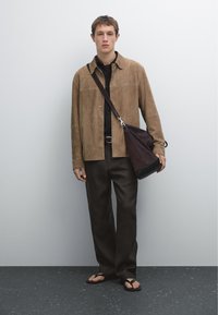 Brown suede button-up shirt, black shirt underneath, dark pants, flip-flops, and a dark bag with silver hardware. Standing against a plain background.
