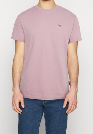 Mand iført en ensfarvet lyse lilla kortærmet t-shirt med lille norsk flaglogo på brystet, kombineret med blå jeans.