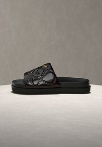 Μαύρο σανδάλι τύπου slide με ανάγλυφο σχέδιο στον ιμάντα και παχιά σόλα, τοποθετημένο πάνω σε ανοιχτή, στρογγυλεμένη επιφάνεια με απλό μπεζ φόντο.