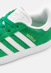 Green suede sneaker with white leather accents and laces. Features three white stripes and "Gazelle" in gold text on the side. Textured sole.
