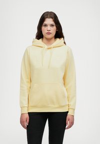 Gul bomullsgenser med frontlomme, snorlukking på hetta og ribbestrikkede mansjetter. Har en liten logo på brystet. Brukes med svarte bukser.