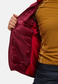 Hand die een kastanjebruine gewatteerde jas vasthoudt, met daarin een rode fleece voering, gedragen over een mosterdkleurig shirt met contrasterende stiksels.