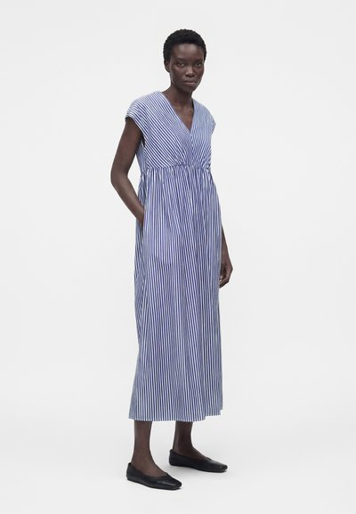 Γυναίκα με μπλε και λευκό κατακόρυφο ριγέ φόρεμα χωρίς μανίκια σε μήκος μέχρι τη μέση της κνήμης, με V λαιμόκοψη και μαύρα φλατ παπούτσια, στέκεται μπροστά σε λευκό φόντο.