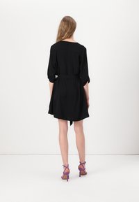 Schwarzes Kleid mit Dreiviertelärmeln, tailliert in der Taille, A-Linien-Silhouette, kombiniert mit lila Riemchenheel. Glatte Textur.