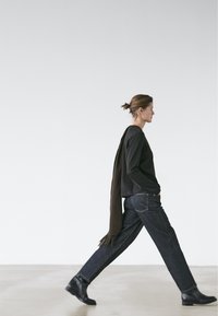 Kvinde går mod højre iført sort langærmet skjorte, mørke jeans, sorte støvler og et langt brunt tørklæde lagt over skulderen.