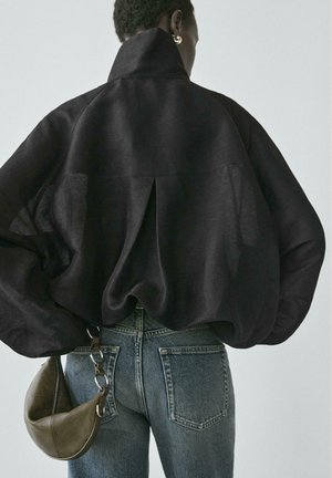 Person trägt eine schwarze übergroße Jacke und blaue Jeans, hält eine braune Ledertasche mit einem silbernen Kettenriemen vor einem einfarbigen Hintergrund.