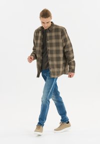 Bruine en beigeruit overhemd met knopen, gedragen over een donkere top, met blauwe spijkerbroek en beige sneakers, op een witte achtergrond.