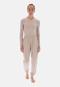 Beżowy długi rękaw i dopasowane spodnie dresowe z falistymi bocznymi akcentami. Miękka tkanina, luźny krój i elastyczna talia. Modelka na boso.