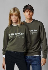 Aliejinės žalios spalvos sportinės palaidinės su įrengtomis rankovėmis. Balta "Red Bull Racing" logotipas. Dėvimos ant juodų ir mėlynų džinsų.