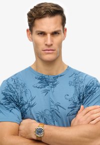 Blue t-shirt with dark tiger and bamboo print; round neck; short sleeves. Brown watch with a blue dial and gold accents on wrist.