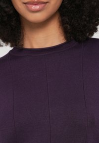 Violets ribota augša ar augstu apaļu apkakli un vertikālām šuvju detaļām. Audums šķiet mīksts ar nelielu mirdzumu.