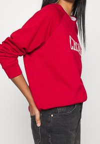 Pull rosso con polsini a coste, scollatura rotonda e testo bianco sul davanti. Indossato con jeans neri, che evidenziano una vestibilità comoda e una consistenza morbida.