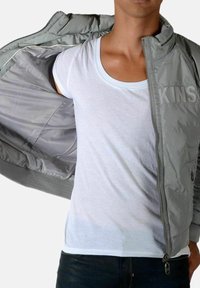 Lysegrå nylon bomberjakke med ribbede manchetter og krave, åbnet for at afsløre en hvid T-shirt under. Klassisk silhuet og tekstur.