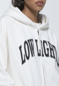 Sudadera blanca con cremallera hecha de tela suave, con un estampado gráfico negro que dice "LOW LIGHT" en el pecho, y con capucha.