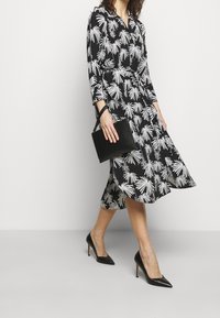 Černé šaty se vzorem bílých palmových listů, dlouhými rukávy, páskem v pase a splývavou sukní. Černá kabelka a černé boty na vysokém podpatku.