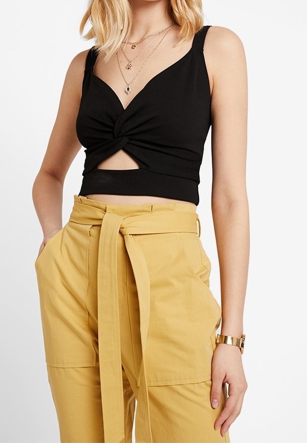 Femme portant un crop top noir sans manches avec un noeud torsadé à l’avant, associé à un pantalon paperbag jaune moutarde taille haute avec une longue ceinture.
