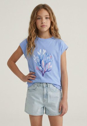 Meisje met lang golvend haar, gekleed in een lichtblauw t-shirt met een kleurrijk koraalontwerp en lichte spijkershorts, staand met één hand op haar heup.