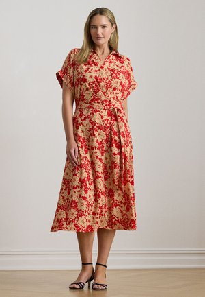 Czerwona i beżowa sukienka kopertowa w kwiaty z krótkimi rękawami, paskiem wiązanym w talii i spódnicą o długości do połowy łydki, wykonana z lekkiego materiału. Czarne sandały na cienkich paskach.