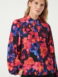 Mulher vestindo uma blusa de mangas longas com um padrão floral abstrato em vermelho brilhante, laranja, azul e roxo, de pé contra um fundo claro e liso.