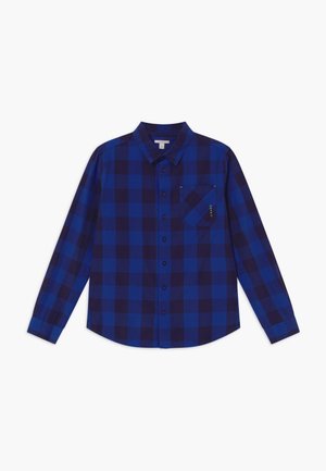 Chemise à manches longues à carreaux bleus et noirs avec col et poche poitrine sur fond blanc.