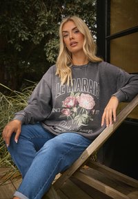 Sudadera gris de gran tamaño con estampado floral y diseño de texto. Jeans de mezclilla azul con puños doblados. Modelo sentado en una silla de madera al aire libre.
