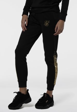 Person iført sorte joggingbukser med guldlogo på venstre lår og guldfoliestribe på ydersiderne, kombineret med sorte sneakers.