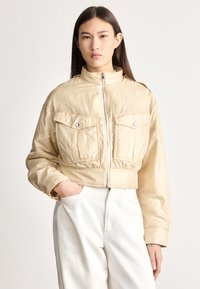 Jauna sieviete bēša krāsas īsā žaketē ar priekšējām kabatām un augstu apkakli, valkājot baltas augsta vidukļa bikses, pozē ar vienu roku kabatā.