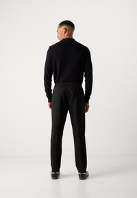 Man standing facing away, wearing a black long-sleeve top, black pants with elastic waistband, and black and white shoes on a white background.