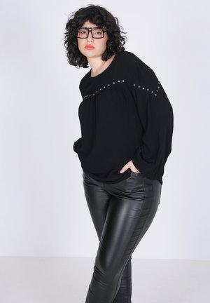 Blouse noire surdimensionnée avec des accents en argent sur les épaules, associée à un pantalon en cuir noir ajusté. Le fond est blanc uni.
