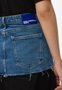 Hátulról látható egy személy, aki kék denim mini szoknyát visel, a Karl Lagerfeld Jeans címkével és rojtos széllel, a keze pedig oldalt látható.