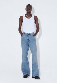 Pantalones de mezclilla de tiro alto y pernera acampanada de color azul claro, con cierre de botones y presillas para cinturón, combinados con una camiseta sin mangas blanca.