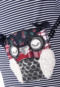 Námořnická a bílá pruhovaná tkanina obsahuje nášivku sovy s květinovými, kostkovanými a flitrovými detaily, včetně texturovaného bílého peří.