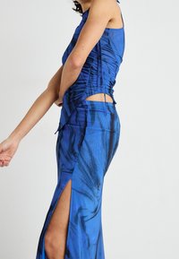 Niebieska sukienka bez rękawów z marmurkowym wzorem, marszczonymi detalami, wycięciami po bokach oraz rozcięciami do wysokości uda, wykonana z gładkiej, zwiewnej tkaniny.