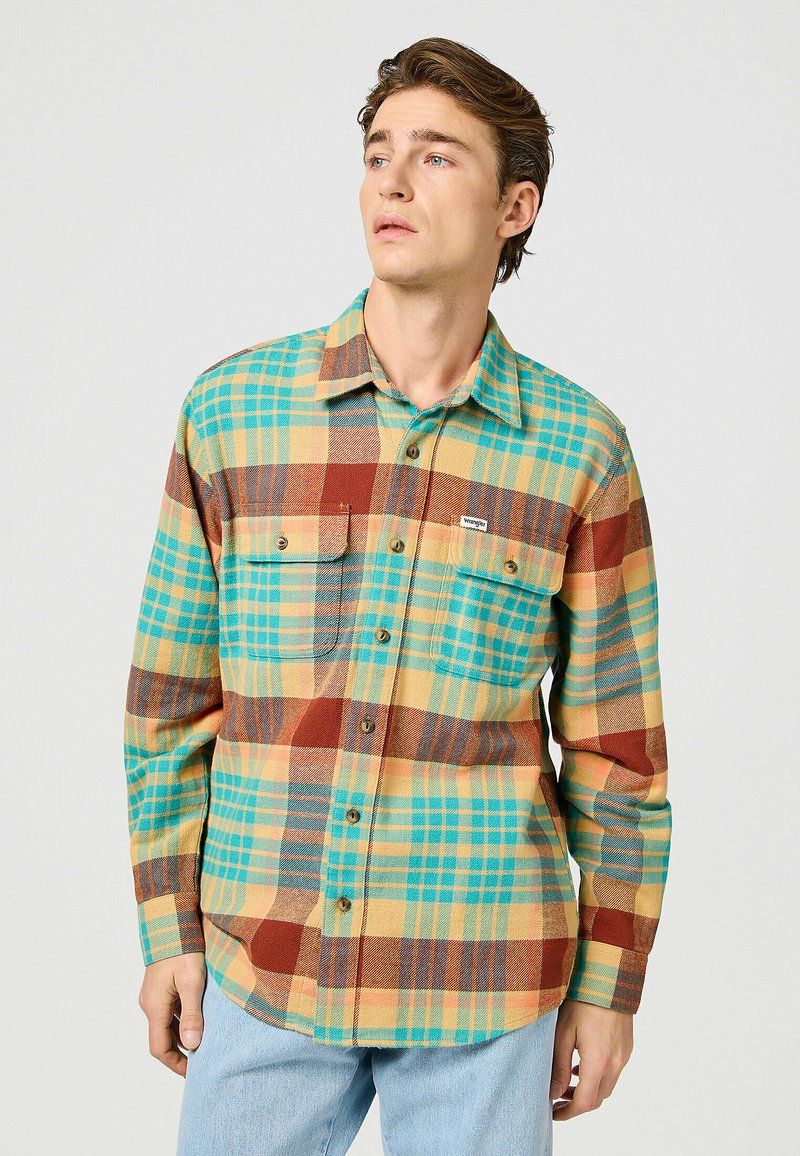 Camisa de franela con un patrón de cuadros multicolor en verde azulado, amarillo, marrón y azul. Confeccionada con un frente abotonado y dos bolsillos en el pecho.