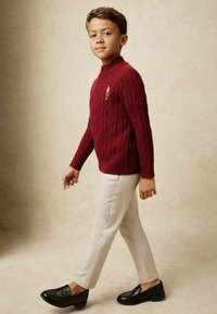 Kootud punane sviiter kõrge kaelusega, millel on nööri muster ja kaunistused. Kombineeritud kreemivärvi sirgete jalgadega pükste ja mustade mokasännidega.