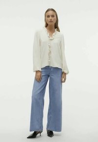 Weiße Bluse mit langen Ärmeln, Rüschenausschnitt und Knopfleiste, kombiniert mit hellblauen weit geschnittenen Jeans und schwarzen High Heels.