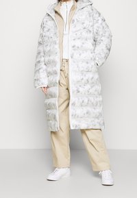 Vadderad kappa i vitt med gråspräckligt mönster, med huva, dragkedjestängning och ribbade ärmar. Kombineras med beige byxor och vita sneakers.