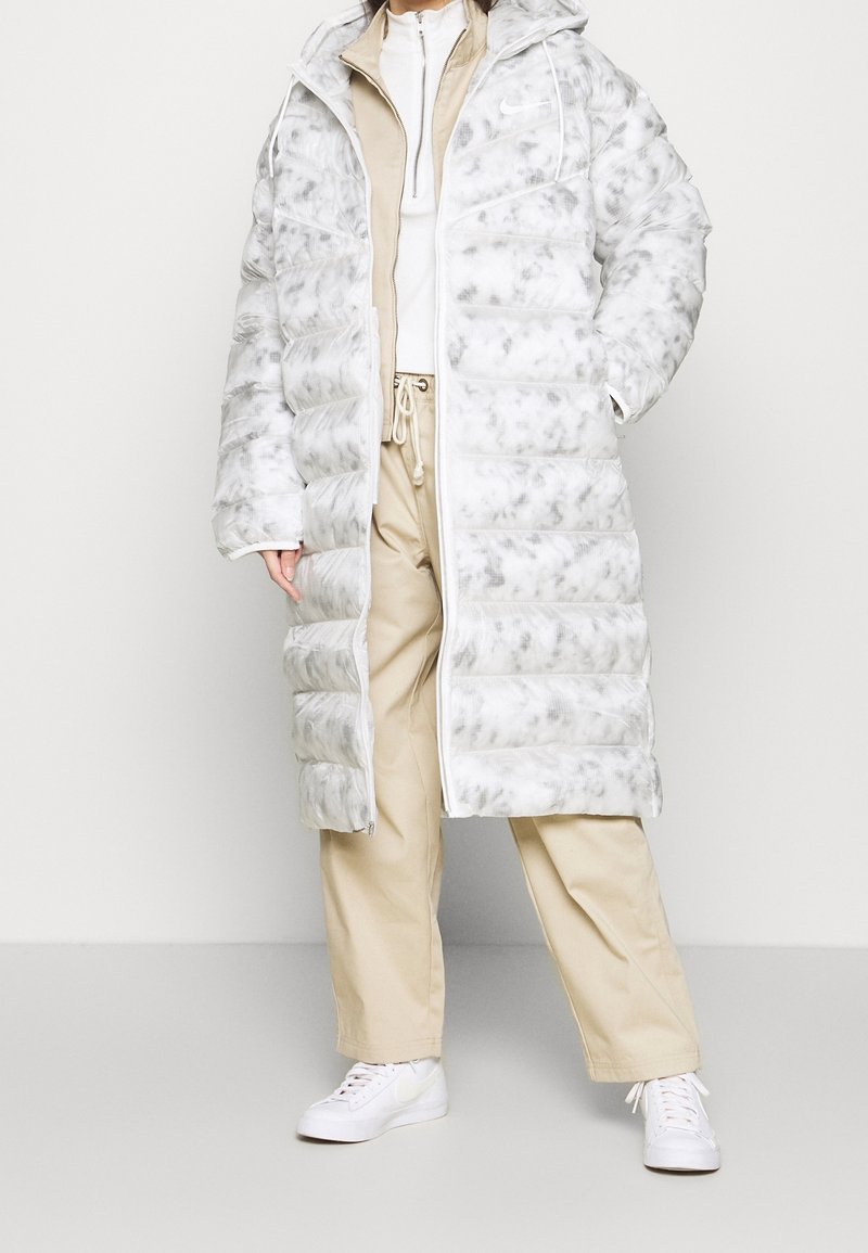 Vadderad kappa i vitt med gråspräckligt mönster, med huva, dragkedjestängning och ribbade ärmar. Kombineras med beige byxor och vita sneakers.