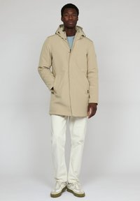 Abrigo impermeable beige con capucha, cierre de cremallera, bolsillos delanteros y un diseño limpio, combinado con pantalones blancos y zapatillas.