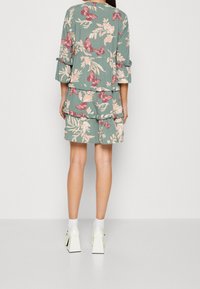 Ensemble floral vert avec un design bouffant, des motifs de papillons dans des nuances de rose et des manches trois-quarts ; associé à des chaussures à talons blocs blanches.